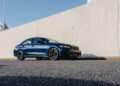 A blue 2026 BMW M340i xDrive sedan is parked on an asphalt road next to a tall, light-colored concrete wall under a clear sky.