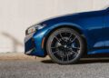 Close-up of the front left wheel and bumper of a 50 Jahre Edition blue BMW M340i xDrive parked on a street, with a beige concrete wall in the background.