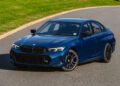 A blue 2026 BMW M340i xDrive sedan is parked on a paved road next to a grassy area under daylight.