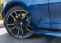 Close-up of a black and silver BMW wheel and tire on a blue 2026 BMW M340i, parked on a sunlit street.
