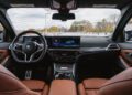 Interior view of a modern 2026 BMW M340i xDrive showing the dashboard, digital displays, steering wheel, and brown leather seats, with a parking lot and trees visible through the windshield.