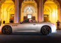A white Maserati convertible sports car with its top down is parked in front of an illuminated building at night, as several people stand nearby taking photos and admiring its Meccanica Lirica elegance.