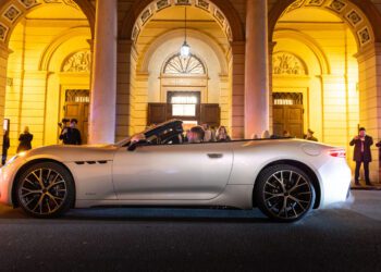 A white Maserati convertible sports car with its top down is parked in front of an illuminated building at night, as several people stand nearby taking photos and admiring its Meccanica Lirica elegance.