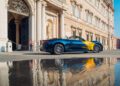 A blue Maserati convertible with yellow detailing is parked outside a grand historic building in Modena, its sleek form elegantly reflected in a puddle on the street.