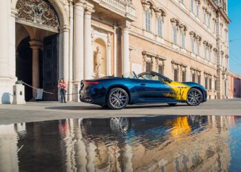 A blue Maserati convertible with yellow detailing is parked outside a grand historic building in Modena, its sleek form elegantly reflected in a puddle on the street.