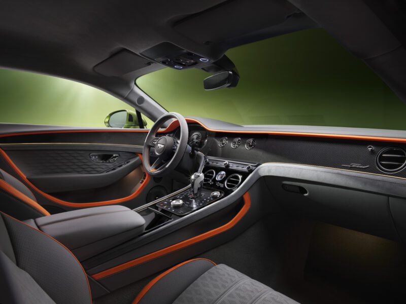 Interior view of a luxury car, reminiscent of the Review: 2026 Bentley Continental GT Speed, featuring black and orange trim, leather seats, a central touchscreen, and a sleek steering wheel and dashboard set against a green background.