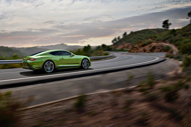 A green sports car, reminiscent of the 2026 Bentley Continental GT Speed, drives quickly along a winding mountain road at sunset, with blurred surroundings indicating motion.
