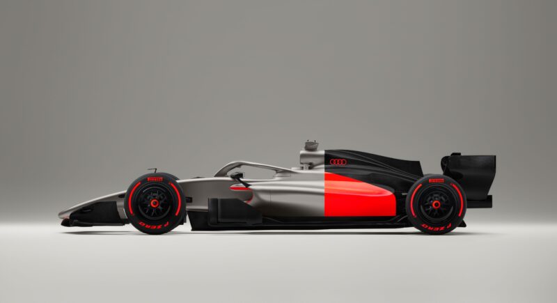 The Audi R26 Concept Formula 1 race car, with a matte silver, black, and red livery and striking red wheel accents, is displayed in a studio setting with a neutral background.