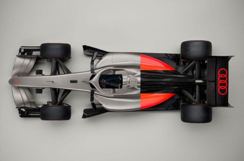 Top view of a sleek, silver Formula 1 Audi R26 concept car with black and orange accents and an Audi logo on the rear wing, photographed on a plain gray background.