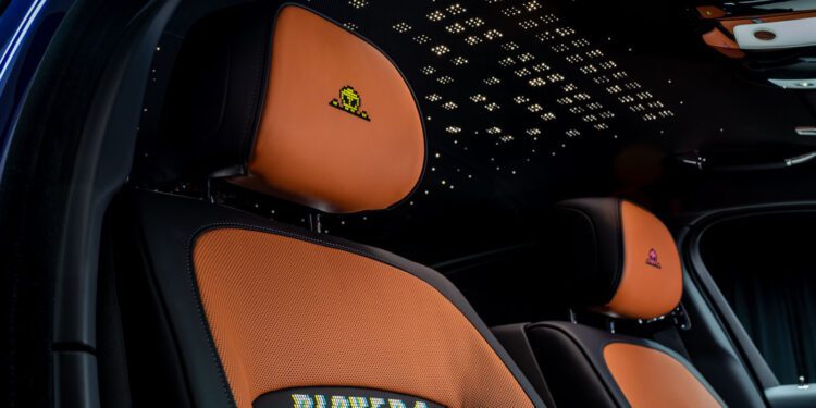 Close-up of two car seats with orange upholstery, embroidered Space Invader icons on the headrests, and "PLAYER 1" and "PLAYER 2" text on the seat backs; starry ceiling above.