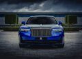 Front view of a blue Rolls-Royce Black Badge Ghost parked on a stone driveway, with dramatic clouds overhead and manicured greenery in the background—a scene fit for any luxury-loving gamer.