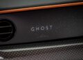 Close-up of a Rolls-Royce Black Badge Ghost dashboard with "GHOST" illuminated among small LED lights, featuring a circular air vent and orange-stitched trim.