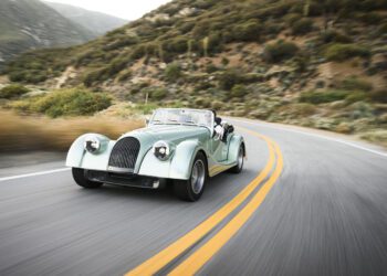 A classic mint green Morgan Plus Four convertible car drives along a winding mountain road lined with vegetation, celebrating the 75th Anniversary of this iconic model in the US.
