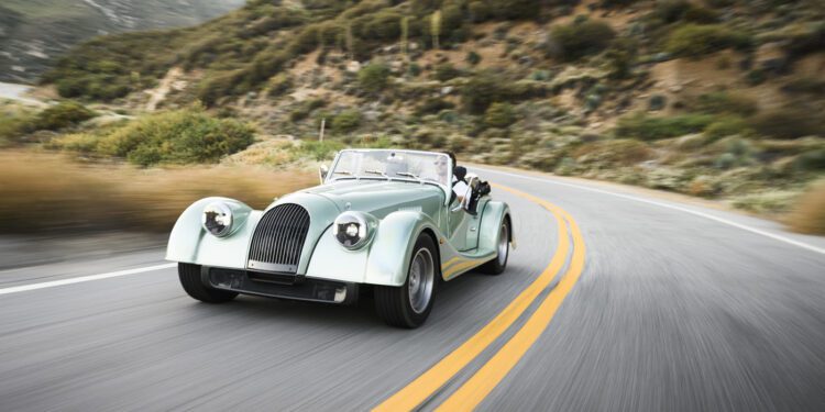 A classic mint green Morgan Plus Four convertible car drives along a winding mountain road lined with vegetation, celebrating the 75th Anniversary of this iconic model in the US.