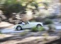 A person drives a 75th Anniversary Morgan Plus Four convertible at speed along a US road, with blurred trees and foliage in the background.