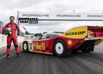 A person in a red racing suit stands next to a vintage Porsche 962 C with Shell and Dunlop branding on the track at Mantrop Park, beneath a Porsche sign.