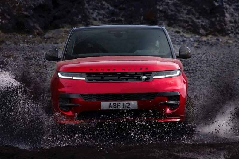 A red Range Rover Sport Dynamic SE PHEV drives through shallow water, splashing up around its wheels, with a rocky landscape in the background. Review: 2025 Land Rover Range Rover Sport Dynamic SE PHEV showcases thrilling off-road capability.