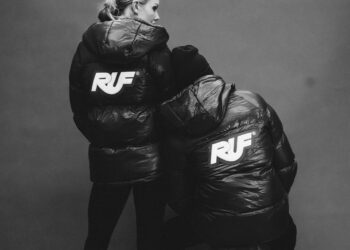 Two people wearing black puffer jackets with large "RUF" logos on the back, from the 2025 Holiday Collection by Aloisa Ruf, stand facing away from the camera against a plain background.
