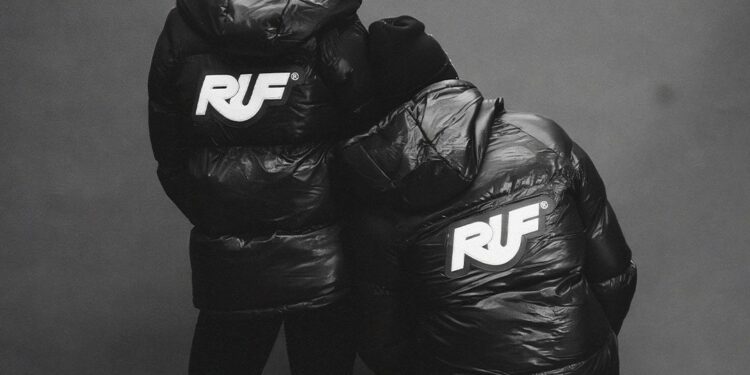 Two people wearing black puffer jackets with large "RUF" logos on the back, from the 2025 Holiday Collection by Aloisa Ruf, stand facing away from the camera against a plain background.