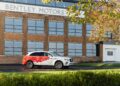 A white Bentley SUV with red graphics, inspired by the Bentley Honors Wartime Heroes with the Czech Squadron Collection, is parked in front of a Bentley Motors building with large windows and brick walls.