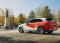 A white and red SUV featuring a heart and lines graphic, inspired by the Bentley Honors Wartime Heroes with the Czech Squadron Collection, is parked near a statue on a paved area, with trees and buildings in the background.