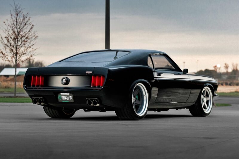 A black classic Ford Mustang fastback, inspired by the Ringbrothers Unveils 800-Plus Horsepower 1969 Ford Mustang Mach 1 'KINGPIN' at 2025 SEMA, features custom wheels and is parked on a paved surface near an empty landscape, seen from the rear left angle.