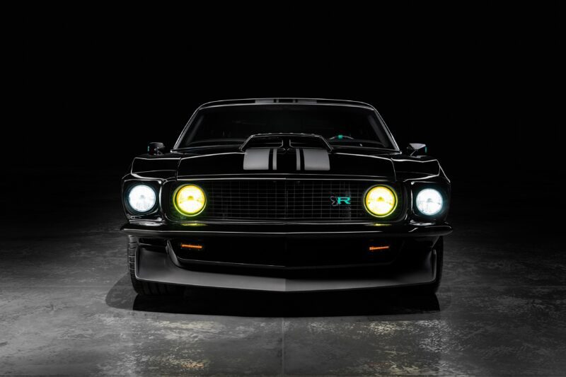 Front view of a black classic muscle car with illuminated headlights, reminiscent of the Ringbrothers' 800-plus horsepower 1969 Ford Mustang Mach 1 'KINGPIN,' unveiled at the 2025 SEMA show, parked on a concrete floor against a black background.