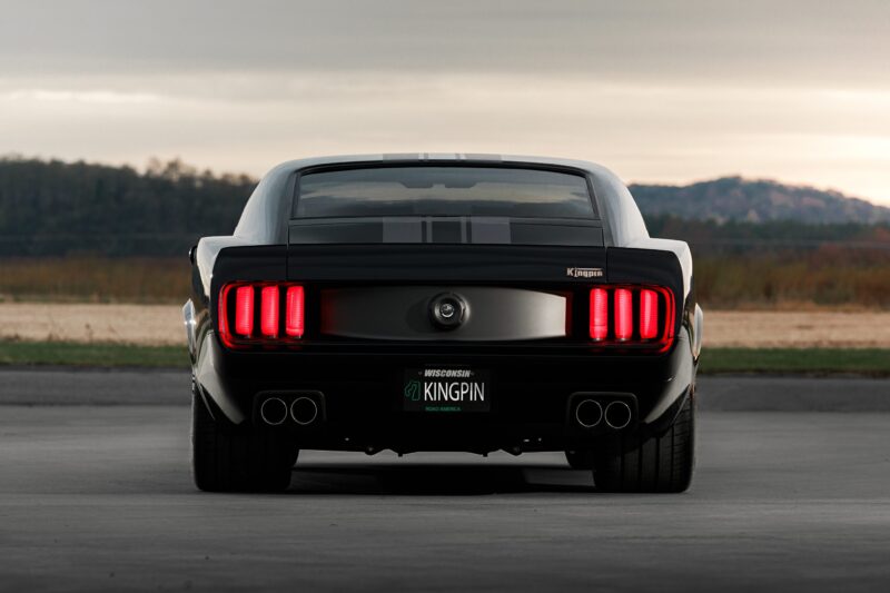A black classic Ford Mustang is parked outdoors, viewed from the rear, with red taillights illuminated and a custom license plate reading "KINGPIN," inspired by Ringbrothers’ 800-plus horsepower 1969 Mustang Mach 1 'KINGPIN' unveiled at 2025 SEMA.