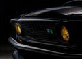 Close-up of the front grille and headlights of a classic black car, reminiscent of the Ringbrothers Unveils 800-Plus Horsepower 1969 Ford Mustang Mach 1 'KINGPIN' at 2025 SEMA, featuring yellow-tinted lights and a blue "R" emblem on the grille.