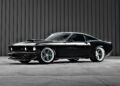 A black classic Ford Mustang with custom modifications, reminiscent of the Ringbrothers' 800-plus horsepower 1969 Mach 1 'KINGPIN' unveiled at the 2025 SEMA, is parked in front of a gray corrugated metal wall.