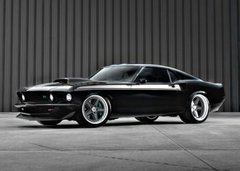 A black classic Ford Mustang with custom modifications, reminiscent of the Ringbrothers' 800-plus horsepower 1969 Mach 1 'KINGPIN' unveiled at the 2025 SEMA, is parked in front of a gray corrugated metal wall.