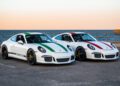 Two white Porsche 911 sports cars—one with green stripes, the other with red—parked side by side by a seaside promenade at sunset, showcase the allure of collector-grade Porsches from Ryan Friedman Motor Cars.