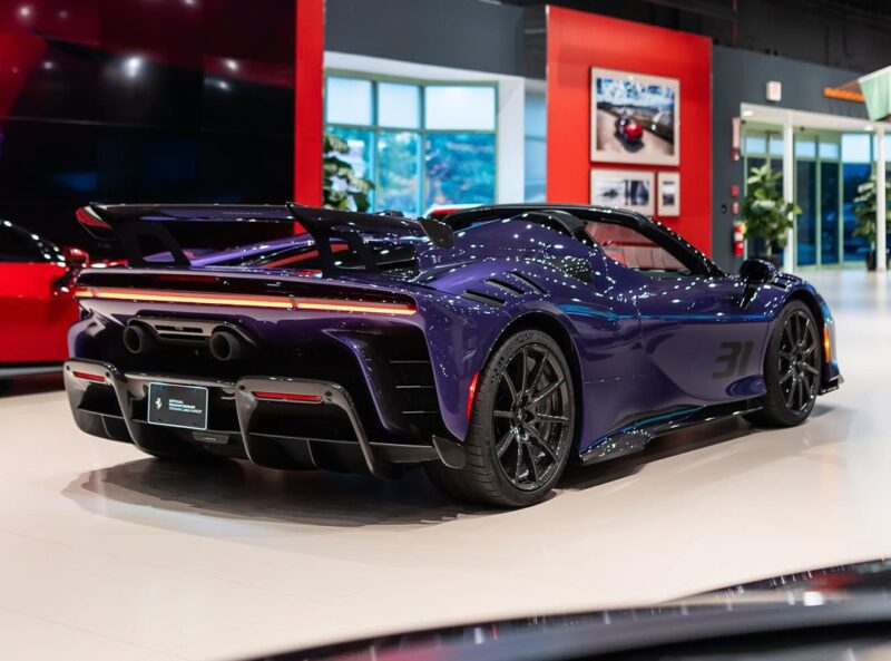 A Violetto Dino SF90 XX Spider is showcased indoors at Ferrari Lake Forest, surrounded by other stunning vehicles and large windows in the background.