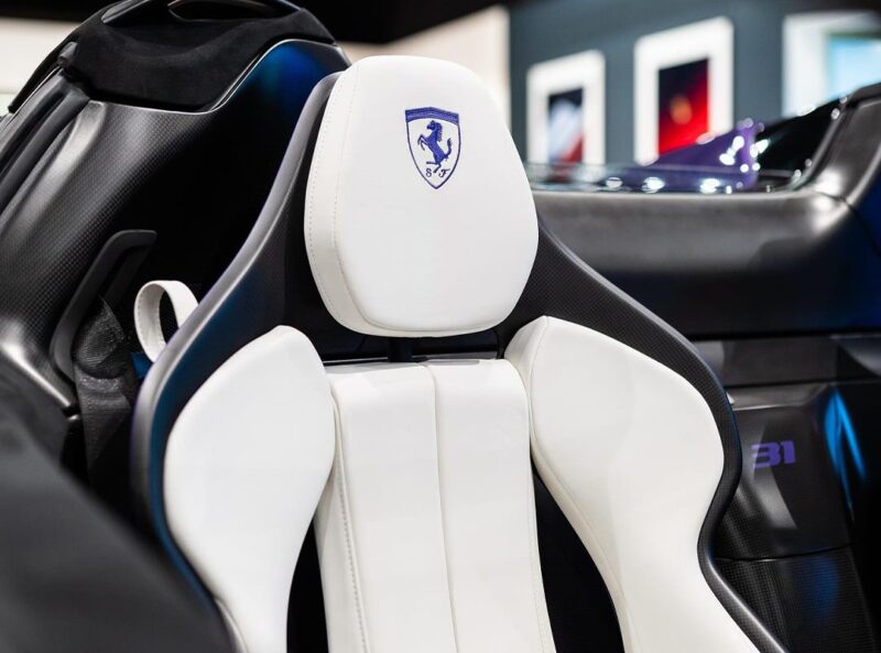 Close-up of a white leather car seat with a blue Ferrari logo on the headrest in an SF90 XX Spider, showcasing part of the car's black and carbon fiber interior at Ferrari Lake Forest.