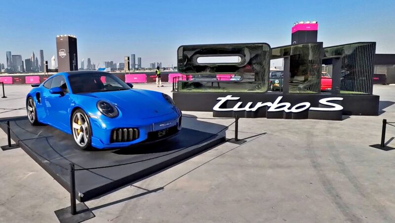 Feature Exclusive: 2025 Icons of Porsche Dubai—a blue Porsche 911 Turbo S is displayed on a platform outdoors, with a bold "911 Turbo S" sign and the city skyline visible in the distance.