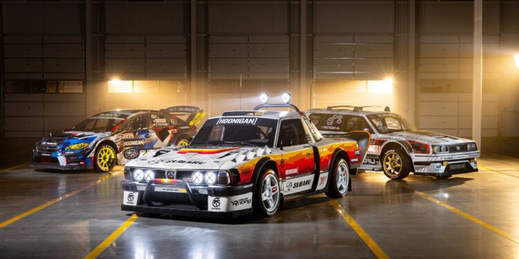 Three modified race cars, including the Subaru Brataroo, with decals and sponsor logos are parked indoors in a well-lit garage with a dark background, showcasing their Gymkhana-ready style for SEMA 2025.