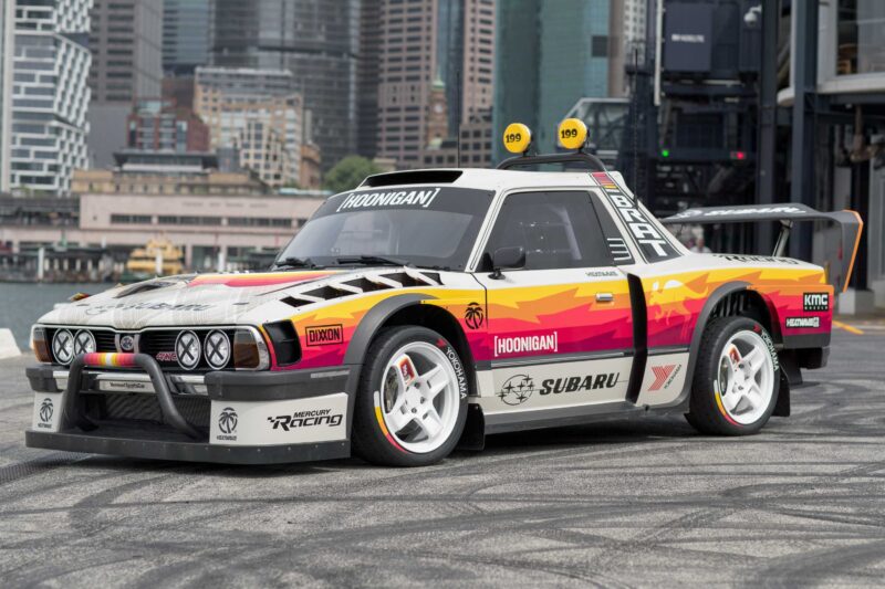 A modified Subaru Brataroo with a wide body kit, racing livery, and rear wing sits on pavement in an urban setting with skyscrapers in the background, ready for Gymkhana action or a SEMA 2025 showcase.