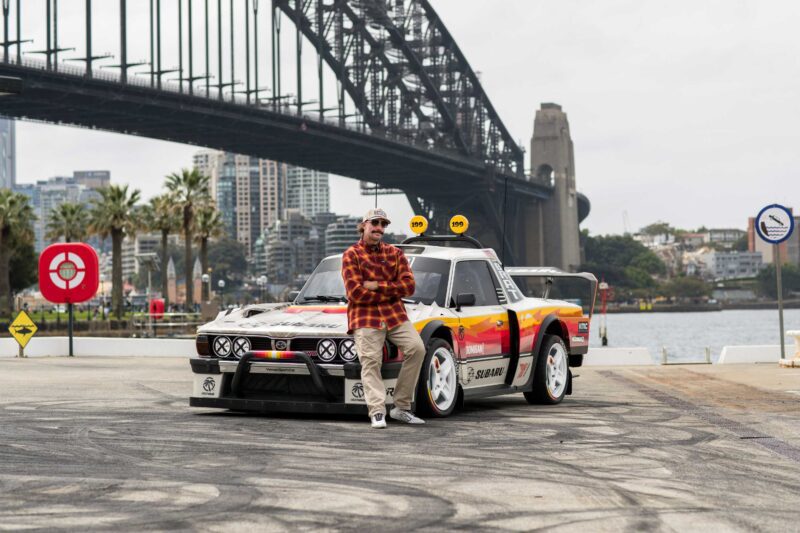 A person in casual clothes leans against a Subaru Brataroo rally car parked near a waterfront, with a large bridge and city buildings in the background—capturing that Gymkhana energy.