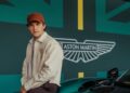 A person wearing a beige jacket and red cap sits on a green Aston Martin car in front of a wall featuring the Aston Martin logo, a stylized British flag design, and the name Tom Holland.