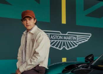 A person wearing a beige jacket and red cap sits on a green Aston Martin car in front of a wall featuring the Aston Martin logo, a stylized British flag design, and the name Tom Holland.