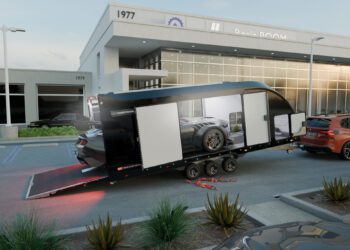 A black Brian James Trailers race transporter with open side and rear doors is parked outside a modern dealership, revealing a car inside. Tools and equipment are scattered nearby.