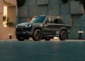 A dark-colored Heritage Customs Defender Pickup is parked outdoors in front of a modern building with geometric concrete panels and minimal landscaping.