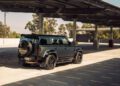 A dark green Custom Defender SUV is parked in a mostly empty, sunlit parking lot under a large metal canopy with trees visible in the background.