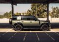 A green SUV with black wheels, possibly a Custom Defender, parked in a covered urban automotive lot, with sunlight casting shadows on the ground.