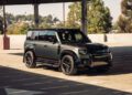 A black Land Rover Defender is parked in a sunlit area of a covered parking structure, blending urban automotive style with trees and buildings visible in the background.