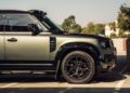 A matte green Defender SUV with black accents is parked in a sunlit outdoor parking lot, showing the front half of the vehicle.