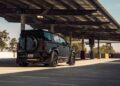 A Custom Cullinan SUV by Urban Automotive is parked under a large solar panel structure in an empty outdoor parking lot on a sunny day, capturing the spirit of SEMA innovation.