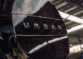 Close-up of a shiny black spare tire cover with the word "URBAN" on it, mounted on the back of a Custom Defender under a metal canopy.