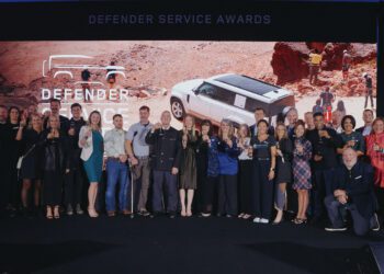 A large group of people pose for a photo on stage in front of a Defender Service Awards sign and an image of a white SUV in a desert landscape.