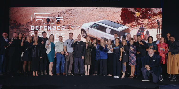 A large group of people pose for a photo on stage in front of a Defender Service Awards sign and an image of a white SUV in a desert landscape.